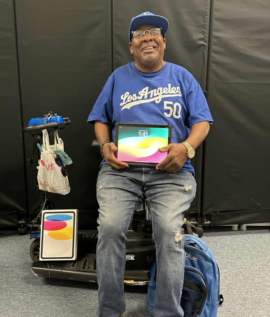 A Rolling Start Inc consumer in a power wheelchair is smiling while holding his brand new iPad. His display screen is on and he shows it towards the camera.